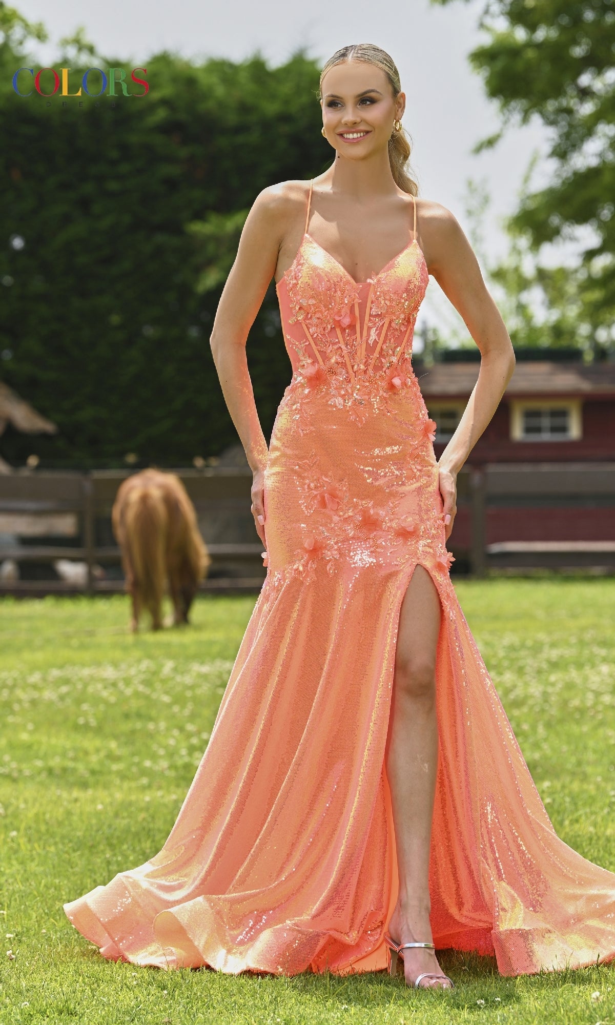 A woman wears the Colors Dress 3540, a long metallic mermaid prom dress with a thigh-high slit, while standing on grass with a horse and barn in the background.